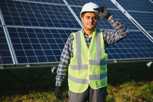 Portrait of Young indian man technician wearing white hard hat standing near solar panels against blue sky. Industrial worker solar system installation, renewable green energy generation concept