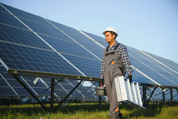 Portrait of Young indian man technician wearing white hard hat standing near solar panels against blue sky. Industrial worker solar system installation, renewable green energy generation concept