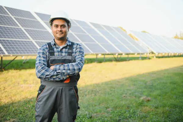 Indian man in uniform on solar farm. Competent energy engineer controlling work of photovoltaic cells.