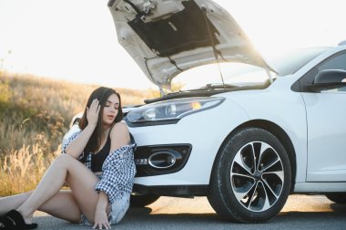 Genç bir kız yolda kırık bir arabanın yanında açık bir kaputla oturuyor.
