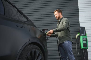 Bir adam şarj istasyonunun yanında durur ve elektrikli arabasını şarj eder..
