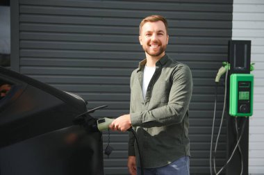 Sıradan giyimli, şarj istasyonunda duran ve elektrikli bir araba için şarj aletinin fişini tutan yakışıklı bir adamın tam boy portresi. Eko elektrikli araba konsepti