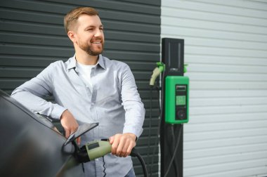 Sıradan giyimli, şarj istasyonunda duran ve elektrikli bir araba için şarj aletinin fişini tutan yakışıklı bir adamın tam boy portresi. Eko elektrikli araba konsepti