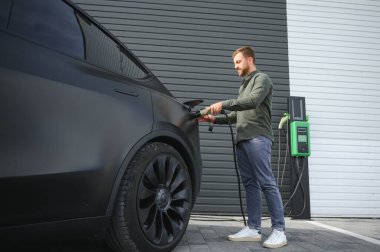 Sıradan giyimli, şarj istasyonunda duran ve elektrikli bir araba için şarj aletinin fişini tutan yakışıklı bir adamın tam boy portresi. Eko elektrikli araba konsepti