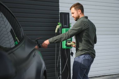 Sıradan giyimli, şarj istasyonunda duran ve elektrikli bir araba için şarj aletinin fişini tutan yakışıklı bir adamın tam boy portresi. Eko elektrikli araba konsepti