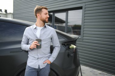 Sıradan giyimli, şarj istasyonunda duran ve elektrikli bir araba için şarj aletinin fişini tutan yakışıklı bir adamın tam boy portresi. Eko elektrikli araba konsepti