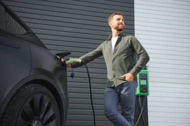 Sıradan giyimli, şarj istasyonunda duran ve elektrikli bir araba için şarj aletinin fişini tutan yakışıklı bir adamın tam boy portresi. Eko elektrikli araba konsepti
