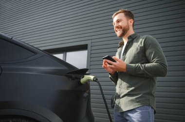 Sıradan giyimli, şarj istasyonunda duran ve elektrikli bir araba için şarj aletinin fişini tutan yakışıklı bir adamın tam boy portresi. Eko elektrikli araba konsepti