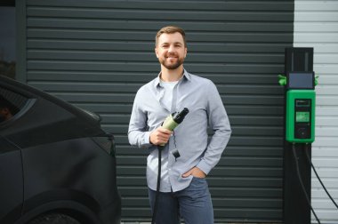 Sıradan giyimli, şarj istasyonunda duran ve elektrikli bir araba için şarj aletinin fişini tutan yakışıklı bir adamın tam boy portresi. Eko elektrikli araba konsepti