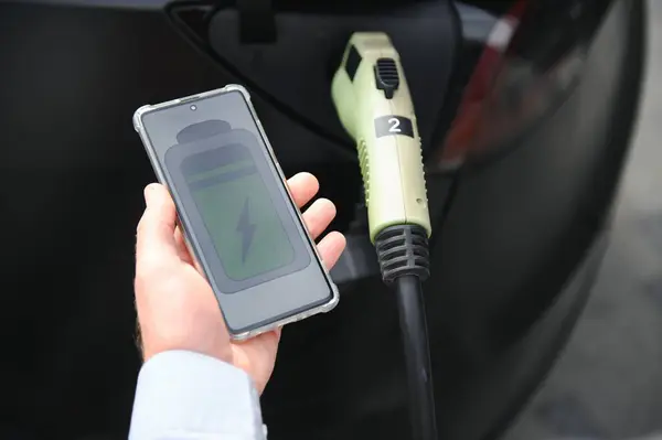 A man charges an electric car and checks the charge level on a smartphone.