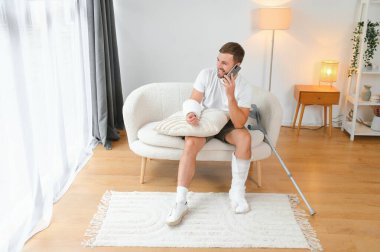 Kırık bacağı ve kolu olan bir adam evdeki kanepede dinlenirken cep telefonunu kullanıyor. Kaza, yaralanma, tedavi, rehabilitasyon kavramı.