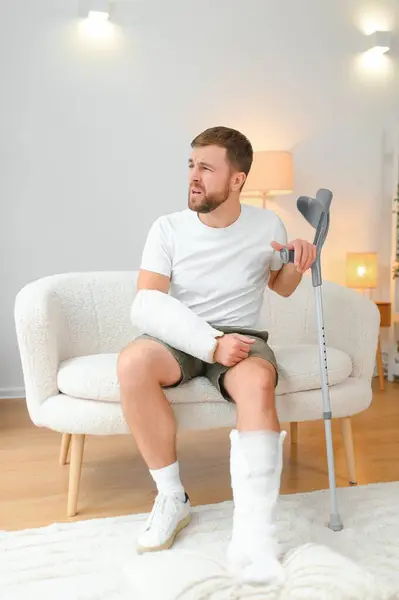 man recovery from accident fracture broken bone injury with leg splints in cast neck splints collar arm splints sling support arm in living room. Social security and health insurance concept.