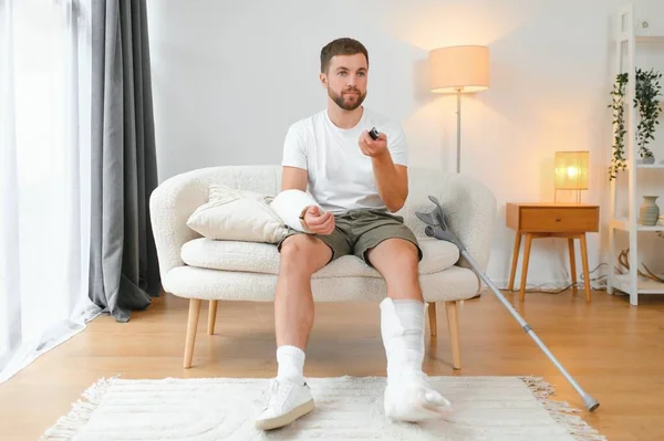 man recovery from accident fracture broken bone injury with leg splints in cast neck splints collar arm splints sling support arm in living room. Social security and health insurance concept.