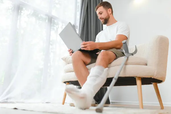 stock image Unhealthy businessman with injured leg and arm in bandage sit on sofa at home work online on computer.