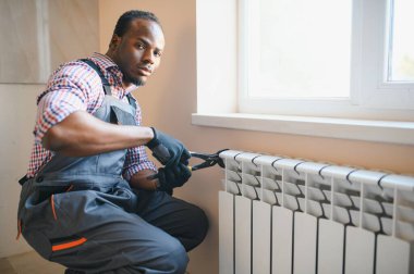 Çalışma kıyafetli bir adam ısıtma kaloriferini yerleştirirken veya tamir ederken araçlar kullanıyor.