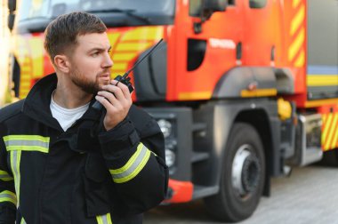 İtfaiyeci üniformalı. Dışarıdaki itfaiye aracının yanında taşınabilir telsiz kullanıyor..
