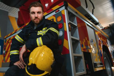 İtfaiyeci üniformalı ve kasklı, itfaiye arabasının yanında..