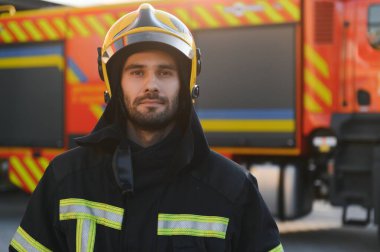 İtfaiyeci üniformalı, istasyonda itfaiye arabasının yanında duruyor..