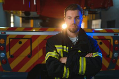 İtfaiyeci üniformalı, istasyonda itfaiye arabasının yanında duruyor..