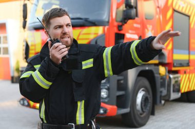 İtfaiyeci üniformalı. Dışarıdaki itfaiye aracının yanında taşınabilir telsiz kullanıyor..