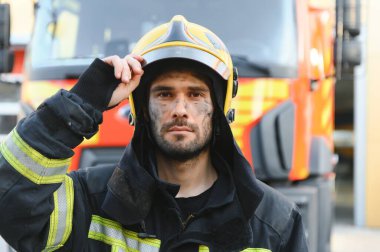 Yangın üzerinde çalıştıktan sonra itfaiye arabasının yanında duran koruyucu bir miğfer ve koruyucu giysili bir itfaiyeci portresi. Resmi kapat..