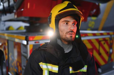 İtfaiyeci üniformalı, istasyonda itfaiye arabasının yanında duruyor..