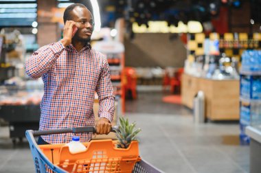 Afro-amerikan erkek müşteri günlük kıyafet içinde alışveriş arabasıyla gülümsüyor ve raflardan yiyecek seçiyor..