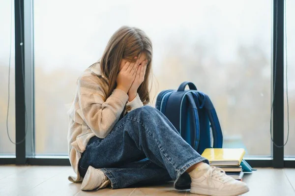 Side view of sad school girl sitting on floor at corridor and hiding face by hands and crying. Concept of hard period of teenagers.