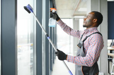 Temizlik işçisi dikkatli ve dikkatli bir şekilde ofis alanının geniş penceresini ovar. Mavi tulumlu ciddi bir Afro-Amerikalı ofisteki çift camlı pencereyi siliyor..