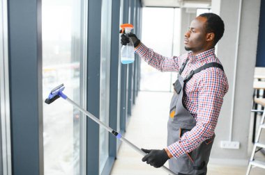 Temizlik işçisi dikkatli ve dikkatli bir şekilde ofis alanının geniş penceresini ovar. Mavi tulumlu ciddi bir Afro-Amerikalı ofisteki çift camlı pencereyi siliyor..
