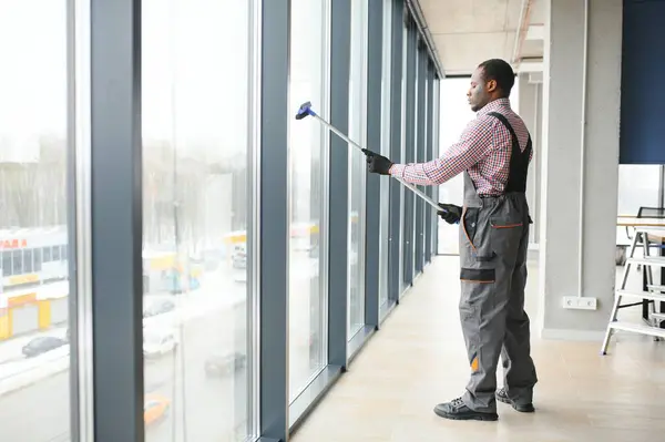 worker of cleaning organization carefully and carefully rubs large window of the office space. A serious African-American in blue overalls wipes the double-glazed window in the office.