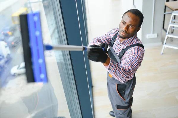 worker of cleaning organization carefully and carefully rubs large window of the office space. A serious African-American in blue overalls wipes the double-glazed window in the office.