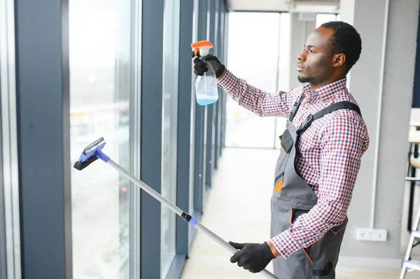 African Male professional cleaning service worker in overalls cleans the windows and shop windows of a store with special equipment.