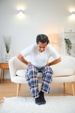 Unhappy indian or arabian man, sits on comfortable sofa in cozy living room, holds his hands on his stomach, grimaces from pain in his stomach, suffers from poisoning, spasm, stomach problems