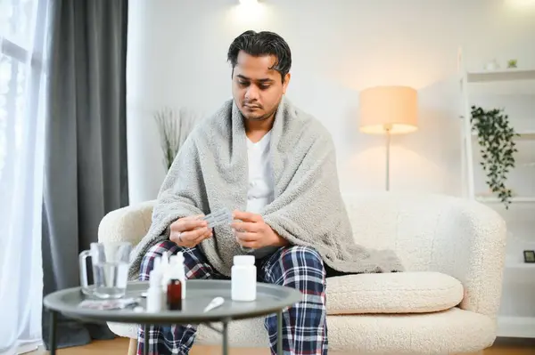 health, cold and people concept - sick young indian man in blanket having headache or fever at home.