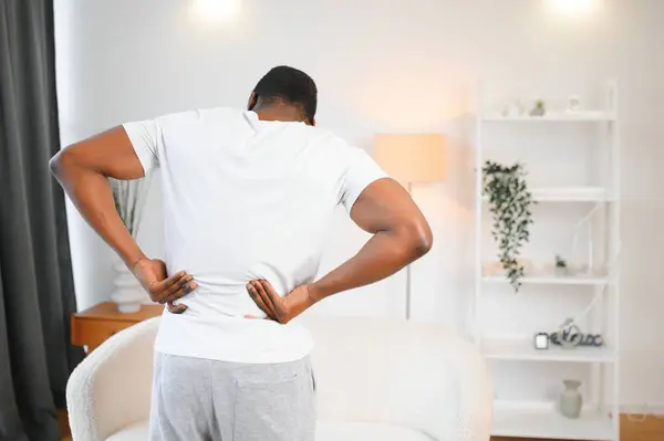 African American guy suffering from back pain, leaning on sofa, cannot walk at home.