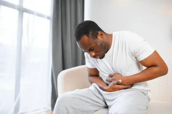 African american guy having stomach ache after eating touching aching stomach suffering from pain sitting on sofa at home. Food poisoning, gastritis, abdominal inflammation and appendicitis.