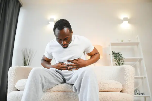 African american guy having stomach ache after eating touching aching stomach suffering from pain sitting on sofa at home. Food poisoning, gastritis, abdominal inflammation and appendicitis.