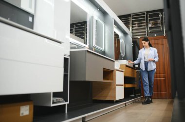 Genç bir kadın, tesisat dükkanında bir sürü hijyenik eşya bulunan yeni banyo mobilyalarını seçiyor..