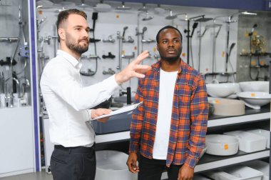 Afrikalı adam ev banyosu için seramik fayans ve kap kacak seçiyor ve erkek satıcı doğru karar vermesine yardımcı oluyor..