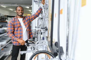 Kahverengi paltolu Afrikalı bir adam inşaat süpermarketinde banyo musluğu seçiyor, evdeki su tesisatını tamir ediyor..