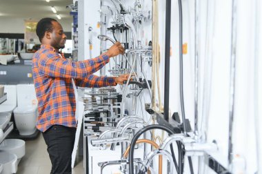 Kahverengi paltolu Afrikalı bir adam inşaat süpermarketinde banyo musluğu seçiyor, evdeki su tesisatını tamir ediyor..