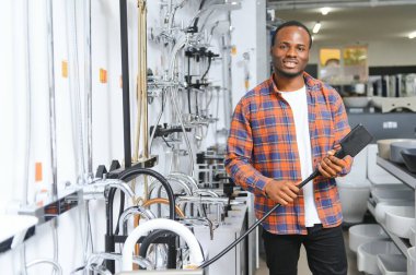 Kahverengi paltolu Afrikalı bir adam inşaat süpermarketinde banyo musluğu seçiyor, evdeki su tesisatını tamir ediyor..