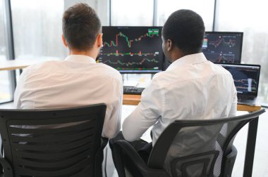 İki farklı iş arkadaşı birbirleriyle konuşuyor, ofiste birden fazla bilgisayar ekranının önünde otururken grafiklere bakıyorlar. Borsa, insanlar, iş konsepti.