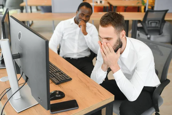 Two Unhappy tired businessman, corporate manager, sits at the workplace, tired from online work, stressed, nervous, massages the bridge of his nose, closed his eyes, experiencing a headache.