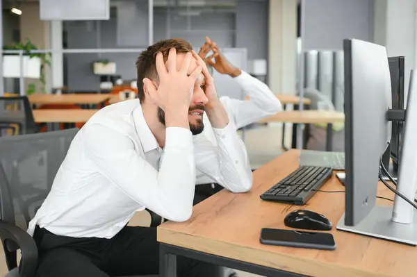 Two Unhappy tired businessman, corporate manager, sits at the workplace, tired from online work, stressed, nervous, massages the bridge of his nose, closed his eyes, experiencing a headache.