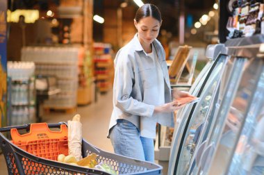 Alışveriş çantası olan genç bir kadın süpermarketteki soğutuculu raftan balık alıyor..