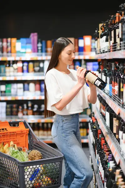 Pretty young woman in casual clothes chooses wine in the supermarket.