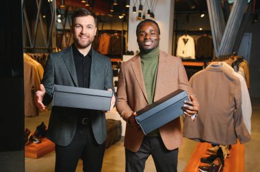 Giysi kutuları taşıyan iki mutlu çok ırklı adam bir giyim mağazasında duruyor..