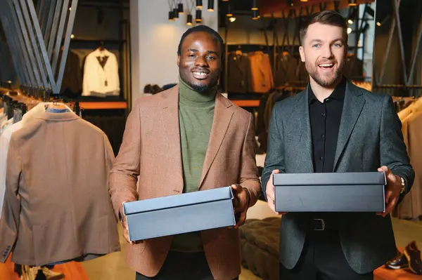 Giysi kutuları taşıyan iki mutlu çok ırklı adam bir giyim mağazasında duruyor..